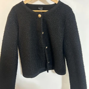 Svart kofta från Gina Tricot - Snygg svart kofta från Gina Tricot med dekorativa guldknappar. Koftan har en texturerad yta och långa ärmar, perfekt för att hålla dig varm och stilren. Passar utmärkt till både vardag och speciella tillfällen.