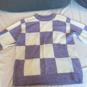 Rutig stickad tröja i lila och vit - Säljer en mysig stickad tröja med ett rutigt mönster i lila och vit. Tröjan har långa ärmar och ribbade kanter vid hals, ärmslut och nederkant. Perfekt för kyliga dagar!