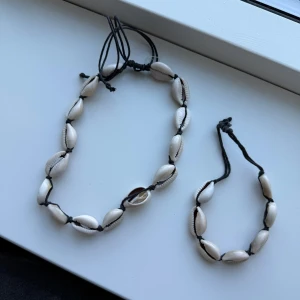Halsband och armband med snäckor - Snyggt set med halsband och armband dekorerade med vita snäckor. Båda smyckena har justerbara svarta snören för perfekt passform. Perfekt för en somrig look!