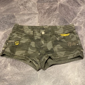 Mini kamouflage shorts med detaljer - Snygga Mini kamouflage shorts med coola broderade detaljer och knappar. Perfekta för en avslappnad stil. De har flera fickor och en knappstängning fram.