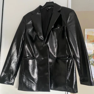 Svart glansig kavaj - Snygg svart kavaj med glansig finish och klassisk krage. Den har en enkel knäppning framtill och två fickor. Perfekt för en stilren look. Är oanvänd. Hör av er om ni vill att jag skickar mer bilder🥰