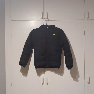 Svart dunjacka från Lyle & Scott - Säljer en svart pufferjacka från Lyle & Scott med deras logga på bröstet. Jackan har långa ärmar och en dragkedja framtill. Perfekt för kyligare dagar. Har en liten skavanker som knappt syns men annars så är den perfekt.