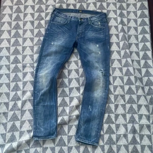 Blå jeans med slitningar - Säljer ett par riktigt snygga blåa jeans med en cool slitningseffekt. De har en klassisk femficksdesign och en bekväm passform. Perfekta för en avslappnad stil.