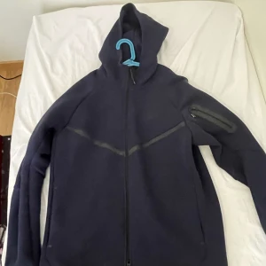 Mörkblå hoodie från Nike - Säljer en mörkblå hoodie från Nike med dragkedja och huva. Jackan har en ficka med dragkedja på ärmen och är perfekt för en sportig look. Den är tillverkad i ett mjukt material som ger komfort och stil. Helt ny och priset diskuteras 