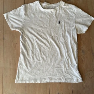 Vit t-shirt från Ralph Lauren - Säljer en klassisk vit t-shirt från Ralph Lauren med det ikoniska broderade logotypen på bröstet. T-shirten har en normal passform och är tillverkad i mjukt bomullsmaterial. Perfekt för en stilren och avslappnad look.