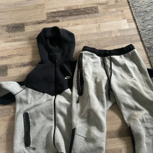Grå och svart nike tracksuit - Säljer en snygg grå och stilren nike tracksuit. Den har en dragkedja framtill och en justerbar huva. Perfekt för en avslappnad stil.