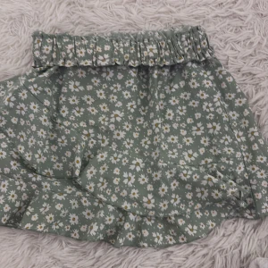 Grön blommig kjol - Söt grön kjol med vita blommor och volangdetaljer. Perfekt för en somrig look med sin lätta och luftiga design. Den elastiska midjan ger en bekväm passform.