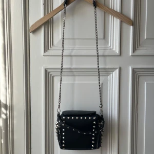 Liten svart axelväska med nitar - Snygg svart axelväska i skinn med coola nitar och kedjeaxelrem. Väskan har en dragkedja och en stilren design som passar perfekt för en edgy look. Perfekt för att ge din outfit en tuff touch.        Använt skick, se bilder, hund finns i hemmet, skicka meddelande för mer info, mått och bilder :) 