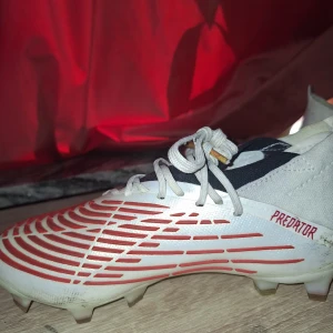 Adidas Predator fotbollsskor - Snygga Adidas Predator fotbollsskor i vitt med röda detaljer och snörning. Perfekta för fotbollsplanen med sin moderna design och funktionalitet.
