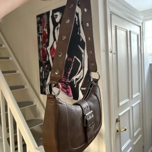 Brun axelväska med spänne - Snygg brun axelväska i skinn med ett coolt spänne framtill. Väskan har en justerbar axelrem med nitar och en dragkedja upptill. Perfekt för att ge din outfit en edgy touch. Prislapp sitter kvar och är köpt för 199 kr.