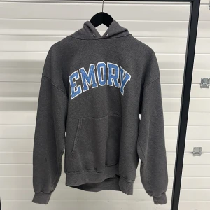 Grå hoodie från Champion - Säljer en grå hoodie från Champion med texten 'Emory' i blått och vitt på framsidan. Tröjan har en klassisk känguruficka och en justerbar huva. Perfekt för en avslappnad stil. Champion-loggan finns broderad på ärmen.