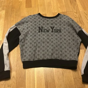 Grå och svart tröja med New York-tryck - Säljer en cool grå och svart tröja med texten 'New York' på framsidan. Tröjan har långa ärmar med vita detaljer och en ribbad kant vid halsen och nederkanten. Perfekt för en avslappnad stil.
