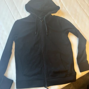 Svart huvtröja från Armani Exchange - Säljer en stilren svart huvtröja från Armani Exchange. Tröjan har dragkedja framtill och en praktisk huva med dragsko. Perfekt för en avslappnad look. Passar även S.