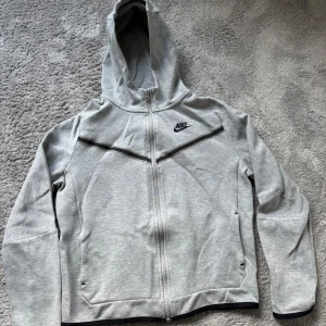 Nike Tech Fleece - Säljer en grå Nike Tech Fleece hoodie med dragkedja och huva.