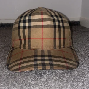 Rutig keps från Burberry - Snygg rutig keps från Burberry i klassiskt beige, svart och rött mönster. Kepsen har justerbar passform med en rem baktill och är perfekt för en stilren look.
