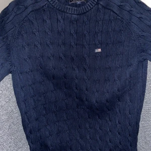 Mörkblå kabelstickad tröja från Lexington - Säljer en stilren mörkblå kabelstickad tröja från Lexington. Tröjan har en liten logotyp på bröstet. Perfekt för en klassisk look.
