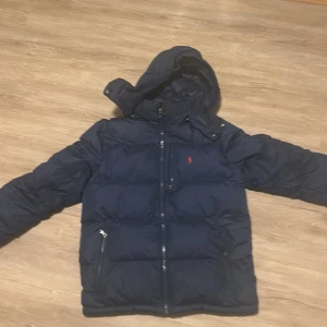 Mörkblå dunjacka från Ralph Lauren - Säljer en snygg mörkblå dunjacka från Ralph Lauren med huva och dragkedja. Jackan har långa ärmar och två praktiska fickor med dragkedjor. Perfekt för kyliga dagar! Köpt för 3000kr  barn jacka storlek 160 cm 