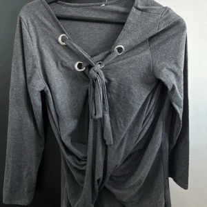 Grå långärmad topp med knytning - Snygg grå långärmad topp med knytning framtill och dekorativa metallringar. Perfekt för en stilren look. Tillverkad i mjukt material för extra komfort.