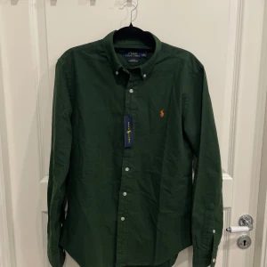 Grön skjorta från Polo Ralph Lauren - Snygg grön skjorta från Polo Ralph Lauren i slim fit. Skjortan har långa ärmar och knappar framtill. Den är tillverkad i stretchigt oxfordmaterial och har en broderad logga på bröstet. Perfekt för en stilren look!