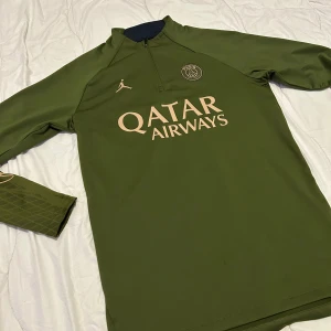 Grön långärmad fotbollströja från Jordan x PSG - Snygg grön långärmad fotbollströja från Jordan i samarbete med Paris Saint-Germain. Tröjan har en halv dragkedja och PSG-loggan på bröstet. Perfekt för fotbollsfans som vill ha stil både på och utanför planen.
