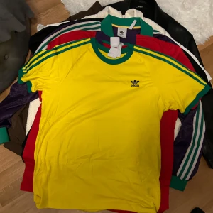 Gul t-shirt från Adidas - Säljer en gul t-shirt från Adidas med gröna och röda ränder på ärmarna och runt halsen. Den har en klassisk Adidas-logga på bröstet. Perfekt för en sportig look!