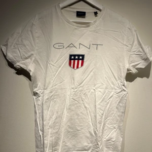 Vit t-shirt från GANT - Säljer en stilren vit t-shirt från GANT med korta ärmar. T-shirten har ett tryck med GANT-loggan och ett sköldemblem i rött, vitt och blått på framsidan. Perfekt för en avslappnad look.