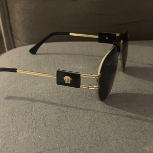 Versace solglasögon  - Snygga svarta solglasögon med guldfärgade detaljer och en ikonisk logotyp på sidorna. De har en klassisk aviatorform som ger en tidlös look. Perfekta för att ge en touch av lyx till din stil.
