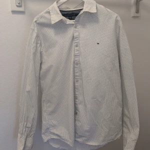 Vit prickig skjorta från Tommy Hilfiger - Säljer en stilren vit skjorta från Tommy Hilfiger med små mörkblå prickar. Skjortan har långa ärmar och knappar framtill. Perfekt för en snygg och avslappnad look.