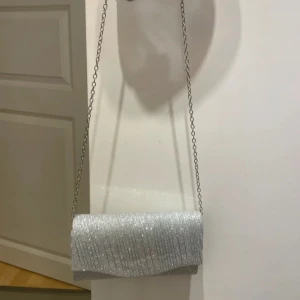 Glittrig silverfärgad kuvertväska med kedja - Snygg och elegant kuvertväska i glittrigt silvermaterial.  Perfekt att använda till ex en bal