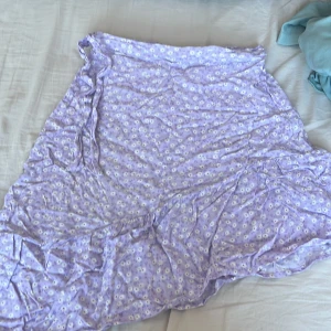 Lila blommig kjol från Kappahl - Söt lila kjol från Kappahl med ett blommigt mönster i vitt. Kjolen har en avslappnad passform och är perfekt för varma dagar. Den är gjord i ett lätt och bekvämt material som gör den skön att bära. Perfekt för sommarens alla äventyr! 🌸