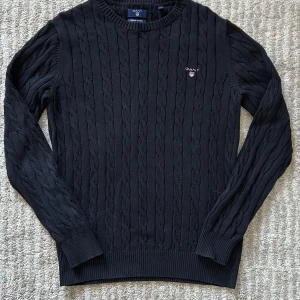 Svart kabelstickad tröja från GANT - Säljer en stilren svart kabelstickad tröja från GANT i premium bomull. Tröjan har en rund halsringning och långa ärmar med ribbade muddar. Perfekt för en klassisk och tidlös look.