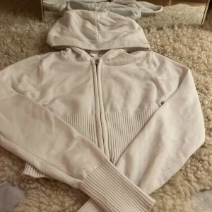 Vit hoodie från Gina Tricot - Säljer en vit hoodie från Gina Tricot med dragkedja och ribbade detaljer vid midjan. Perfekt för en avslappnad stil. Den har en bekväm huva och långa ärmar.