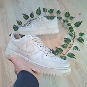 Vita Nike Air Force 1 sneakers - Säljer ett par klassiska vita Nike Air Force sneakers. De har en stilren design med snörning och är tillverkade i skinn. Perfekta för en casual look.
