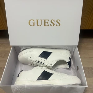 Vita sneakers från Guess - Snygga vita sneakers från Guess med svarta detaljer på sidan. Skorna har snörning och en klassisk design som passar perfekt till vardags. Skorna är helt nya i sin originals förpackning.