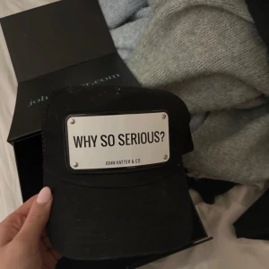 Svart keps från John Hatter & Co - Snygg svart keps med texten 'WHY SO SERIOUS?' på en metallplatta framtill. Perfekt för en cool och avslappnad stil. Kepsen kommer i en elegant förpackning.