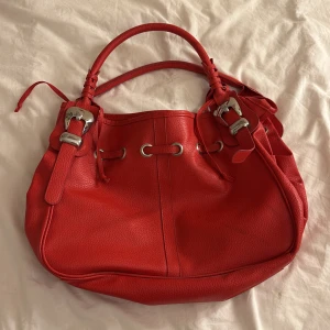 Röd axelväska  - Snygg röd axelväska i skinn med silverfärgade detaljer och dekorativa detaljer. Väskan har två handtag och en stilren design med dragkedja och knytband. ganska stor. har en fläck som jag inte får bort därför säljer jag den billigare 