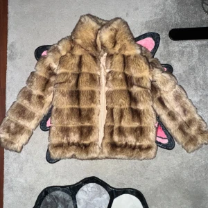 Brun pälsjacka - Säljer en lyxig brun pälsjacka med fluffig textur och hög krage. Jackan har en stilren design utan synliga knappar eller dragkedja på framsidan. Perfekt för att hålla sig varm och trendig under kyliga dagar.