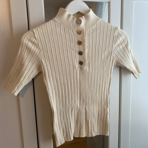 Beige ribbad topp med knappar - Snygg beige ribbad topp med korta ärmar och knappar framtill. Perfekt för en stilren look. Den har en hög krage och är tillverkad i ett mjukt material. Knapparna är dock lite repiga, men det ser man knappt.