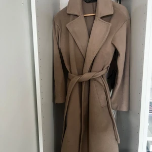 Beige kappa från Amisu - Säljer en stilren beige kappa från Amisu. Den har en klassisk design med knytbälte i midjan och långa ärmar. Perfekt för höst och vår. Kappan har en elegant krage och är tillverkad i ett mjukt material. Storleken är 34 men den är oversize och går att reglera med bandet därav passer den även 36-38