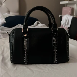 Svart handväska med nitar - Snygg svart handväska i skinn med coola nitar längs kanterna. Den har två fack inuti en med dragkedja och en utan. Aldrig använd i super fint skick. Axelrämsband tillkommer. Köpt för 500kr