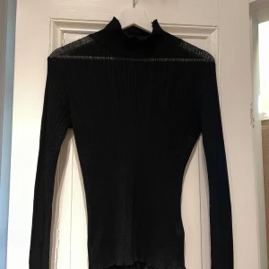 Svart ribbad topp från Josefine HJ - Snygg svart ribbad polotröja från Josefine HJ x na-kd. Tröjan har långa ärmar och en hög krage som ger en stilren look. I ”see through” material.