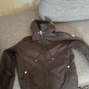 Svart vindbreaker från Moncler - Snygg svart vindjacka från Moncler med dragkedja. Jackan har Moncler-logga på ärmen och detaljer i rött, vitt och blått på huvan. Perfekt för blåsiga dagar. Lite trasig vid moncler märket och på vänster armen( då jag ramlade en gång med jackan). Annars i bra skick. Storlek 2/M men passar 1/S