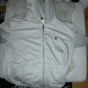 Grå hoodie från Polo Ralph Lauren - Säljer en stilren grå hoodie från Polo Ralph Lauren med dragkedja och det klassiska broderade logotypen på bröstet. Hoodien har en bekväm passform och är perfekt för en avslappnad look. Den har en justerbar huva och praktiska fickor framtill.