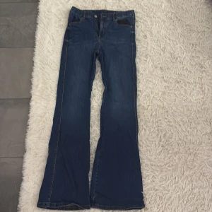 Blå midwaist bootcut jeans - Snygga blå bootcut jeans med klassisk femficksdesign och knappgylf. Perfekta för en avslappnad stil med en touch av retro. Passar bra till både sneakers och stövlar.