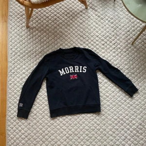 Mörk blå tröja från Morris - Säljer en mörkblå tröja från Morris med vit text och en liten brittisk flagga på bröstet. Tröjan har långa ärmar och en klassisk rund halsringning. Perfekt för en stilren och avslappnad look.