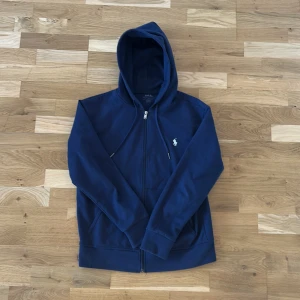 Mörkblå huvtröja från Polo Ralph Lauren - Säljer en snygg mörkblå huvtröja från Polo Ralph Lauren. Tröjan har en dragkedja framtill och en liten broderad logga på bröstet. Perfekt för en avslappnad stil med sin bekväma passform och långa ärmar.