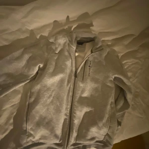 Grå jacka från Sail Racing - Säljer en stilren grå jacka från Sail Racing med dragkedja framtill. Jackan har en hög krage och en liten ficka med dragkedja på bröstet. Perfekt för en casual look.