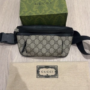 Gucci Midjeväska  - Snygg Gucci magväska med klassiskt GG-monogram. Väskan har en dragkedja och justerbart bälte för perfekt passform. Den är tillverkad i syntetmaterial med svarta detaljer och kommer med originalförpackning. Serienummer stämplad från Gucci ingår på väskan.