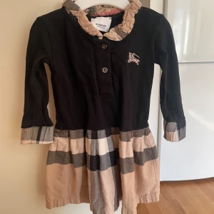 Barn Svart och beige klänning från Burberry - Säljer en söt svart och beige klänning från Burberry med rutigt mönster på kjolen och volangdetaljer vid kragen. Klänningen har långa ärmar och knappar framtill. Perfekt för en stilfull look! Storlek 74/80
