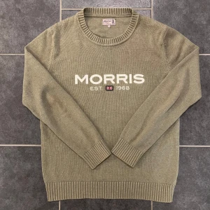 Khakigrön bomullströja från Morris - Säljer en stilren khakigrön tröja från Morris i 100% bomull. Tröjan har en rund halsringning och ribbade muddar. Framtill finns en broderad logotyp med texten 'MORRIS EST. 1968' och en liten flagga. Perfekt för en avslappnad och snygg look.